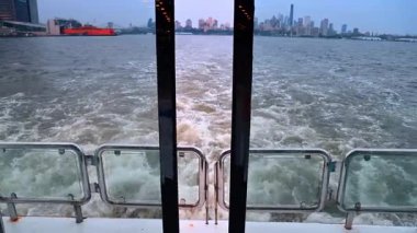 New York Limanı 'nda feribot kalkışı. Feribotun arkasındaki dalgalı su izleri New York Limanı 'ndan şehir siluetine doğru ilerliyor..