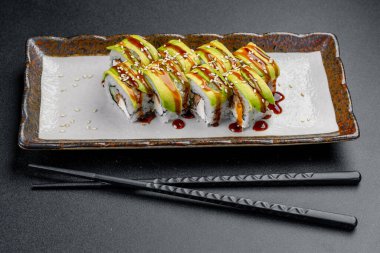 Avokado ile suşi ruloları ve yemek çubuklarıyla servis edilen bir tabakta sos. Avokado ve sosla kaplı suşi ruloları yanında siyah çubuklarla bir tabağa konur..