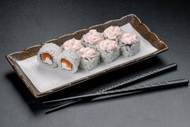 Suşi ruloları ve tabakta pembe tatlılar ve karanlık bir masada yemek çubukları. Balıklı suşi ruloları ve pembe tatlılar karanlık bir yüzeyde yemek çubuklarının yanında bir tabakta otururlar..