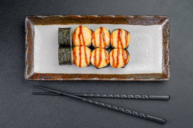 Suşi ruloları, siyah bir masada yemek çubuklarıyla ahşap bir tabağa konur. Suşi dürümleri ahşap bir tabakta, yanında siyah yemek çubukları ile birlikte siyah bir masada güzelce düzenlenir..