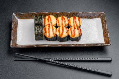 Sos ile servis edilen suşi ruloları ve siyah bir yüzey üzerinde çubuklar. Suşi ruloları, üzerine sos serpiştirilmiş bir tabakta güzelce düzenlenir.