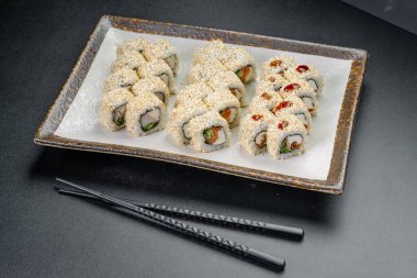 Restoran masasında siyah bir tabakta yemek çubuklarıyla servis edilen suşi ruloları. Bir tabakta suşi ruloları ve susam tohumları ve sos var. Yemek masasında yemek çubuklarıyla yemeye hazır..