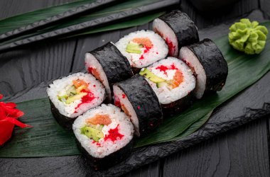 Suşi, yeşil garnitür ve kırmızı çiçeklerle siyah taştan bir tabakta yuvarlanıyor. Suşi ruloları yeşil yapraklı ve kırmızı çiçekli siyah taştan bir tabakta düzenlenir. Wasabi de yakınlarda..
