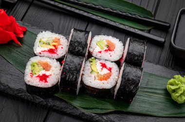 Suşi ruloları yanında çubuklar olan siyah ahşap bir masaya dizilmiş. Suşi ruloları siyah bir yüzeye yerleştirilmiş. Yanında yeşil garnitür ve çubuklar var..