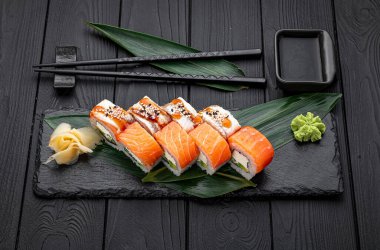 Suşi yuvarlakları, yemek çubukları ve baharatlarla birlikte koyu taştan bir tabağa dizilmiş. Suşi ruloları, yakınındaki yemek çubukları, zencefil, wasabi ve yeşil yapraklarla birlikte koyu bir tabağa konur..