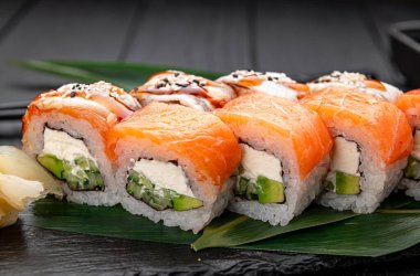 Siyah ahşap bir masada somon ve krem peynirli suşi ruloları. Suşi ruloları yeşil yapraklı siyah ahşap bir masanın üzerine düzgünce dizilir. Somon ruloları kaplar ve renk katar.