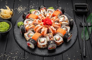 Siyah ahşap bir masada çeşitli rulo ekmeklerle servis edilen suşi tabağı. Büyük bir tabakta birden fazla suşi rulosu bulunur. Suşinin yanına yeşil wasabi ve turşulu zencefil konur..