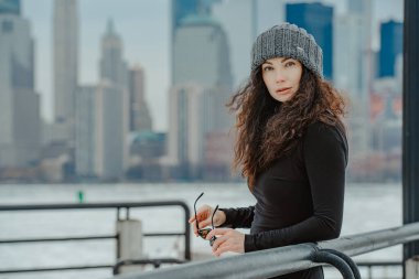 New York silueti geçmişi olan bir kadın portresi. Gri bereli ve siyah bluzlu bir kadın portresi. Arka planda bulanık Manhattan gökdelenleri olan güneş gözlüğü..