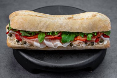 İtalyan ciabatta sandviçi, jambonlu roka ve kapari. Parma jambonlu baget, krem peynirli domates roka, siyah ahşap tezgahta gri desenli arka planda..