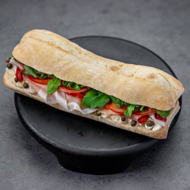 Arugula kapari ve krem peynirli jambonlu sandviç. Parma jambonlu baget sandviç roka kapari siyah ahşap kaidede gri desenli arka planda..