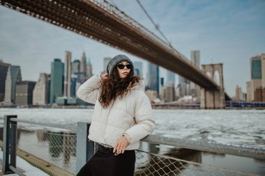Brooklyn Köprüsü siluetine karşı beyaz ceketli kadın. Brooklyn Köprüsü ve Manhattan ufuk çizgisi geçmişi olan Doğu Nehri kıyısında kışlık elbiseli bir kadının portresi..