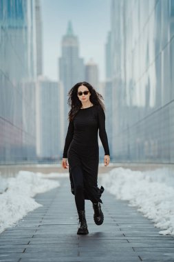 Siyah elbiseli bir kadın New York gökdelenlerinde yürüyor. Manhattan şehir merkezindeki cam ofis binaları arasında karlı bir kaldırımda yürüyen bir kadının tam vücut fotoğrafı..