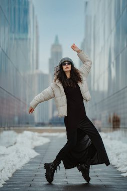 New York 'un kışın gökyüzünde poz veren şık bir kadın. New York 'ta cam binalar arasında poz veren uzun siyah elbiseli ve beyaz ceketli bir kadının tam boy fotoğrafı..