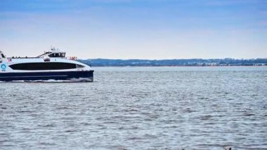 Körfezi geçen Katamaran feribotunun yan görüntüsü. Mavi ve beyaz ticari feribot uzak kıyı şeridi ve arkaplanda ufuk çizgisiyle su üzerinde hareket ediyor..