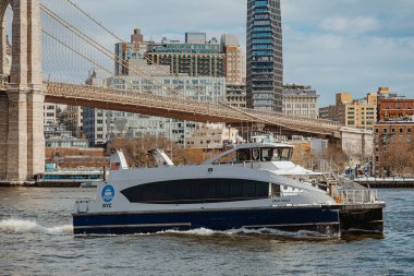 New York, ABD, 23 Şubat 2026: Brooklyn Köprüsü 'nün altından geçen feribot. East River 'daki yolcu feribotunun yan görüntüsü. Şehir binaları ve köprü yapısı var.