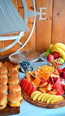 Ahşap yemek tepsisindeki mini çizburger dilimleri. Birbirinin aynısı pek çok kürdanlı mini burger, deniz duvarı dekorasyonuna karşı ahşap bir tahtanın üzerinde servis ediliyor..