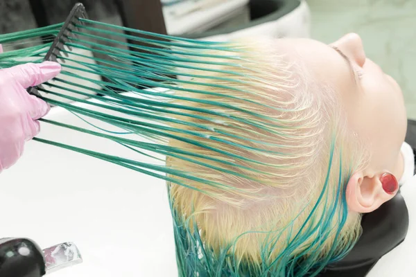 Professional hairstylist in protective glove holds of hair in hand and brushes long green and bleached hair of client while washing head in shower