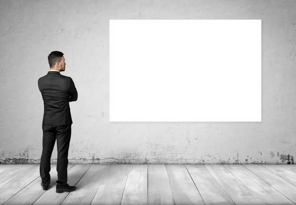 Young man in business suit, standing front of concrete wall with white blank board on it. — Stock fotografie