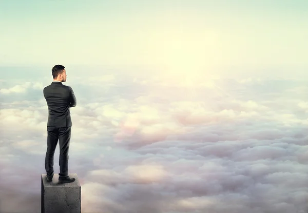 Young man in business suit, standing on concrete column above clouds. — Stockfoto
