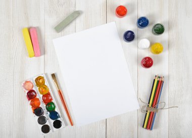 Work place of designer with colored pencils, brush, gouache jars, watercolor paints, chalks and a white paper in top view clipart