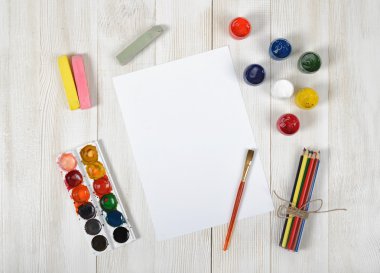 Work place of designer with colored pencils, brush, gouache jars, watercolor paints, chalks and a white paper in top view clipart