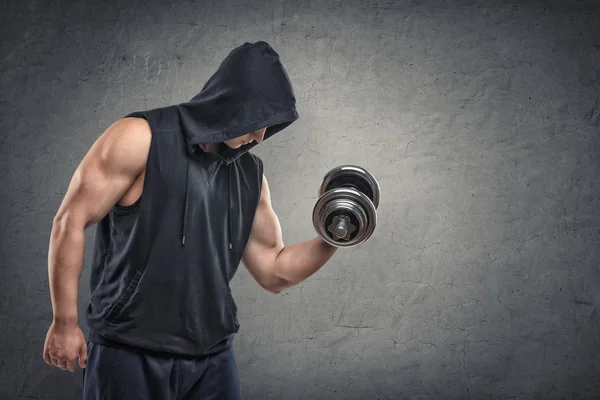 Muscular young guy in hoodie lifting a dumbbell to show his biceps — Stock Photo, Image