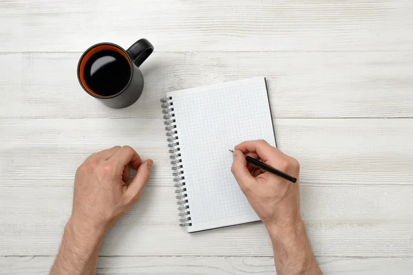 Hands of man writing with pencil in notebook and cup coffee standing on wooden table — Stock Photo, Image