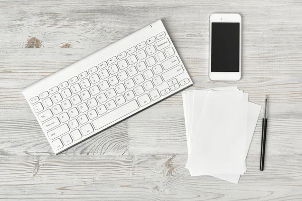 Pracoviště s klávesnicí, smarthphone, bílé listy a pera na dřevěné desce v pohledu shora. — Stock fotografie