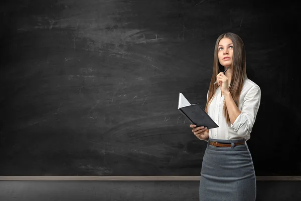 Young woman with notebook and pen looking pensively on the background of blackboard — Stock Fotó