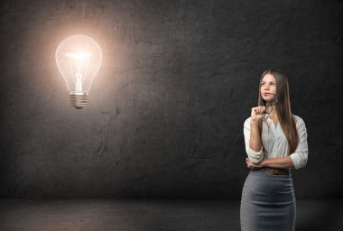Young businesswoman with glasses looking thoughtfully on the glowing light bulb