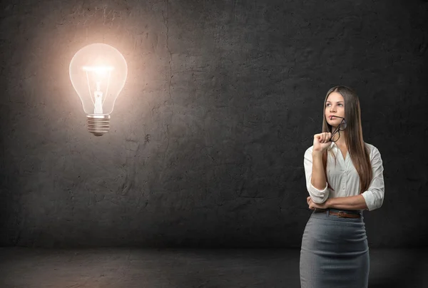 Young businesswoman with glasses looking thoughtfully on the glowing light bulb — Zdjęcie stockowe