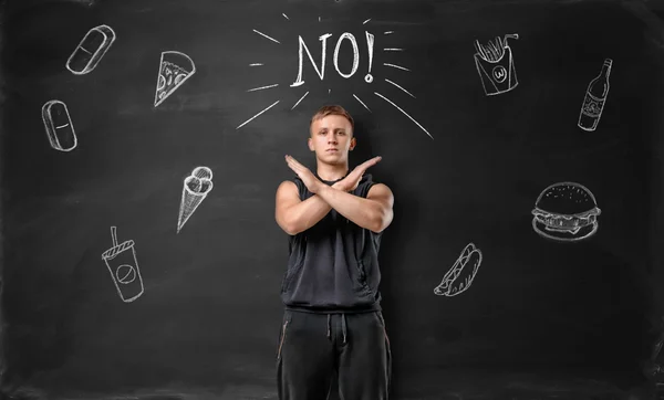 Muscled young man showing stop sign with his hands and says no to unhealthy food on the background of blackboard — Stock Photo, Image