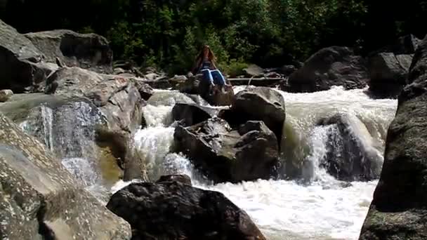 Молодая девушка туристка сидит и брызгает водой на камне среди реки — стоковое видео
