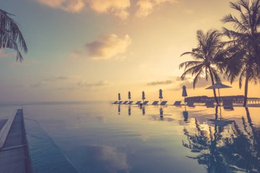 Günbatımı havuz kenarında su yansıması manzarası. Havuzu, plaj sandalyeleri ya da şemsiyeleri altında palmiye ağaçları ve günbatımı gökyüzü olan lüks plaj tatil köyü. Yaz seyahati ve tatil geçmişi kavramı. Lüks sonsuz havuz