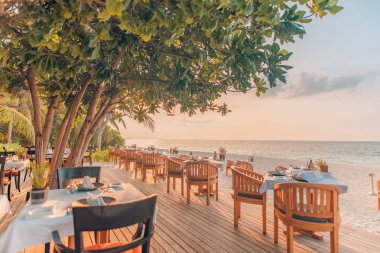 Outdoor restaurant on island resort beach. Table dinner setting at the beach on twilight time. Amazing romantic dinner on the beach on wooden deck under sunset sky. Romance and love, luxury destination dinning, exotic table setup with sea view