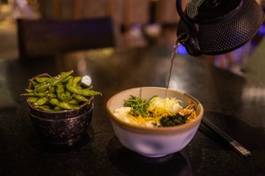 Çaydanlık, bir kase eriştenin yanına koyu bir masada bir kase edamame döker..