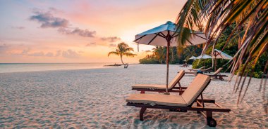 Plajda gün batımı, şezlonglar ve yumuşak turuncu ve pembe gökyüzünün altında bir şemsiye..