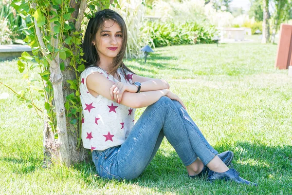 Junge brünette Frau sitzt auf dem Rasen unter einem Baum — Stockfoto