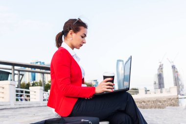 Kırmızı elbiseli bir iş kadını deniz kenarındaki otoparkta dizüstü bilgisayarla oturuyor. Serbest Çalışan.