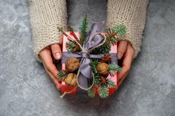 Noel hediyesi, kırmızı süsleme, kadın ellerinde beyaz örgü süveter, gri arka planda çam kozalakları ve Noel süslemeleri.