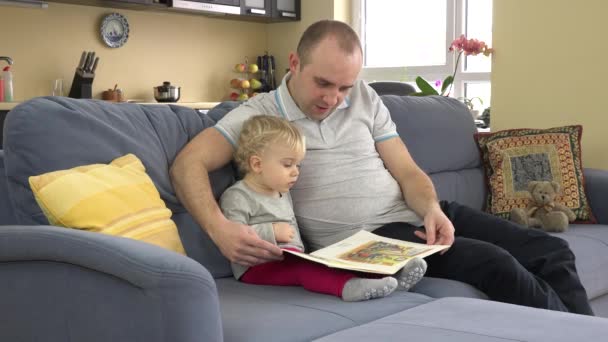 Roztomilá dívka s mladým otcem podívat se na obrázky v knize doma na pohovce. — Stock video