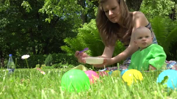 A mãe com colher alimenta o purê de legumes do bebê perto de plantas de samambaia no parque. 4K — Vídeo de Stock