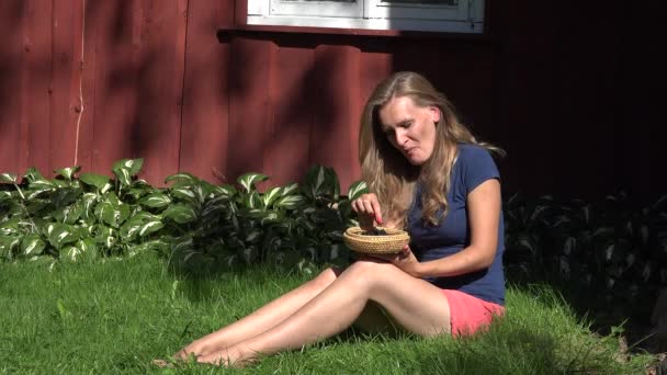 Mulher feliz em shorts sentado perto da casa de madeira rural comendo amora preta e sorriso. 4K — Vídeo de Stock