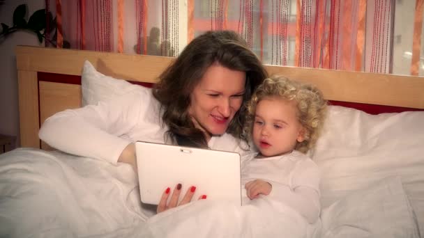 Mutlu anne ve kıvırcık kız yatakta tablette çizgi film izliyorlar. 4K — Stok video