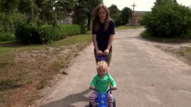 Anne, yaz zamanı doğada üç tekerlekli bisiklete binen küçük bir kız taşıyor. 4K