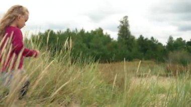 Deniz kenarındaki uzun otların arasında kum tepelerinde yürüyen aktif bir kız. 5 yaşında bir çocuk