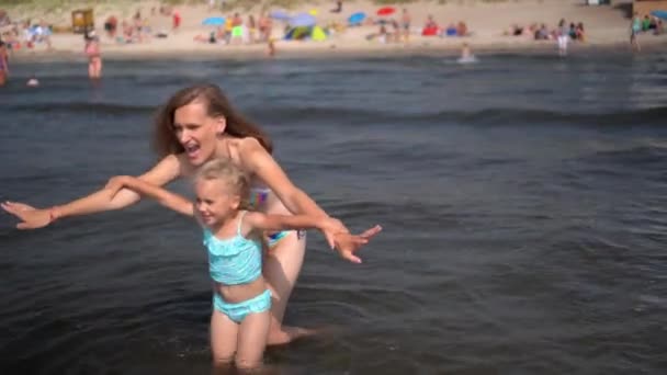 Verspielte Mutter und ihre kleine Tochter vor den Wellen des Meeres. Verschwommener Strand mit Menschen — Stockvideo