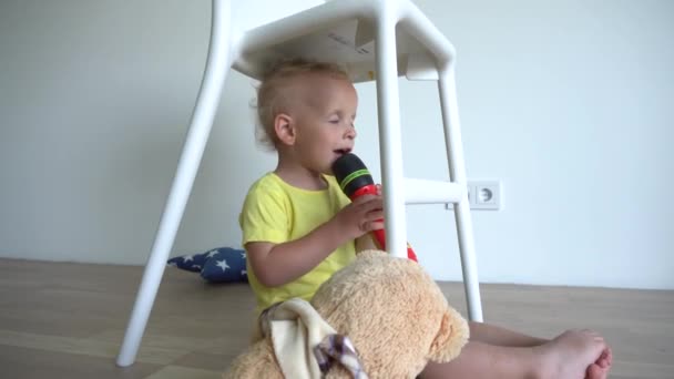 Menino feliz cantar com microfone de brinquedo sentado debaixo da cadeira. Loira criança se divertir — Vídeo de Stock