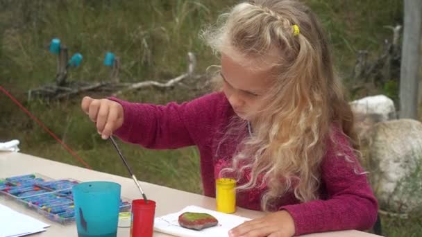 Маленька дівчинка дитина з гуашем і пензлем розмальовує камінці, що сидять за столом — стокове відео