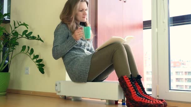 Woman read book, drink warm tee sitting on radiator in room. 4K — Stock Video